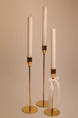 Three white wax candles stand in elegant golden candlesticks. The stem of one of the candlesticks is decorated with a white ribbon bow.