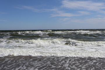 Powerful ocean waves crash onto a sandy beach, creating a foamy shoreline against a backdrop of clear blue skies. A serene yet dynamic scene of nature's raw beauty