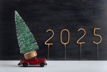 Toy car carries miniature Christmas tree, golden number 2025 on black background. Delivery of...