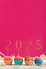 New Years background. Holiday cupcakes with numbers 2025 surrounded by New Years tinsel on pink...