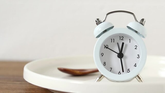 Alarm clock on empty plate and wooden spoon with white background. Intermittent fasting and meal planning concept. Close-up.