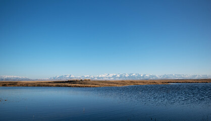 contrasting landscapes where the stillness of water bodies foreground the rugged majesty of distant, snow-capped mountains, framed by an expansive blue sky