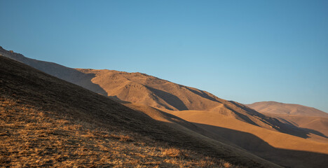 Golden hour light casting long shadows over undulating hills and dry grassland in a serene, high-altitude mountain landscape.