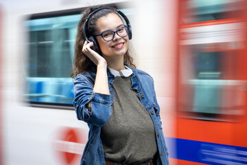 Pretty young girl listening to music with headphones at the train station and looks at the camera