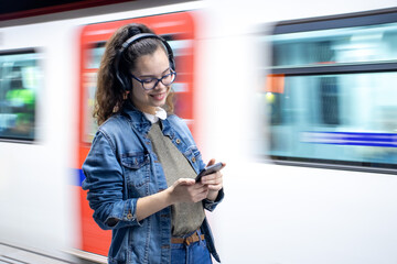 Pretty young girl listening to music with headphones at the train station while looking at her phone