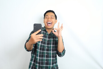 carefree asian man wearing casual plaid shirt laughing and show okay gesture while holding smartphone isolated on white background