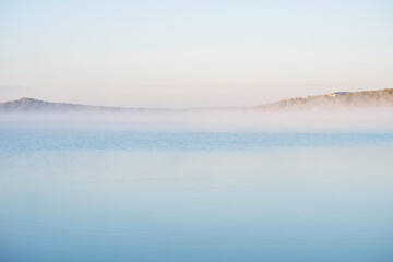 Fototapeta premium a tranquil lake under a soft, pastel sky at dawn or dusk. Mist hovers over the calm water, partially obscuring the tree-lined far shore with hints of autumnal colors