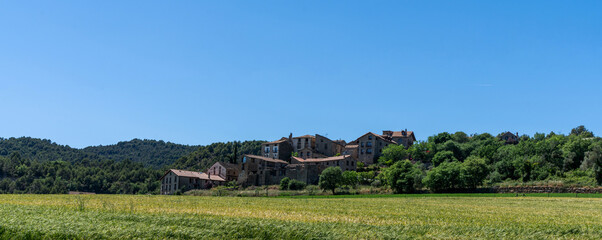 A golden sea of wheat sways gently in the breeze, foregrounding a cluster of stone houses nestled among verdant trees, with the calm presence of hills rising in the background under a clear sky.