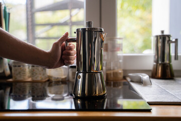 Woman preparing coffee at home kitchen setting near window, girl holding a modern stainless geyser coffee pot on induction stove panel indoor, geyser coffee maker