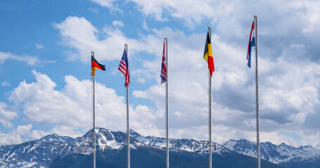 National flags of different countries flying in the wind against a blue sky and snowy mountain peaks.