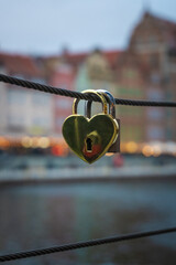 A golden heart-shaped padlock hangs from a bridge railing, symbolizing love and commitment. In the background, the cityscape and river appear softly blurred, enhancing the romantic atmosphere.