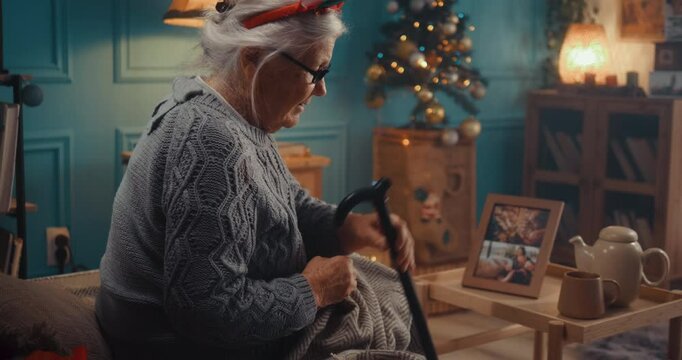The old grandmother celebrates Christmas alone at home, feels sad and lonely sitting on the bed, and looks at family photos. Sad retired senior 90s woman who misses her family during winter holidays