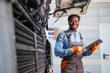 Black truck mechanic or maintenance engineer standing by the vehicle and holding tablet computer inside workshop.