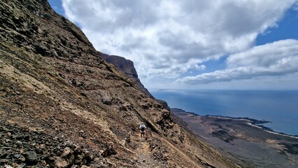 A hiker navigates a rugged mountain trail along a dramatic cliffside overlooking the expansive sea under a sky dotted with clouds. The scene captures the beauty and challenge of outdoor exploration.