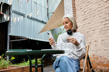 A beautiful woman with gray hair relaxes at a cafe, sipping coffee and using her smartphone.