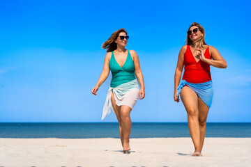 Two beautiful young females wearing one-piece bathing suits and pareo walking and talking on white...