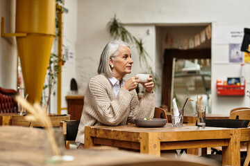A beautiful mature woman with gray hair sits thoughtfully in a warm cafe, sipping her drink.