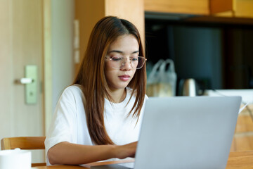 Young Asian woman in glasses focusing on her laptop, dressed casually in a white shirt, in a quiet home environment, with a peaceful expression, working at a wooden table with a coffee cup nearby.