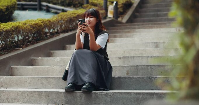 University student, girl and break with phone on stairs for social media scroll, reading ebook or campus website. Japanese, person and relax on steps at college with smartphone for streaming service