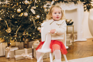 Baby girl riding rocking horse near christmas tree with presents