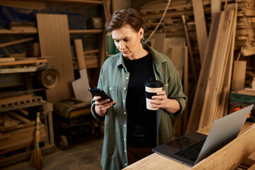 A skilled carpenter examines her smartphone while holding a coffee cup, surrounded by woodworking tools and materials.