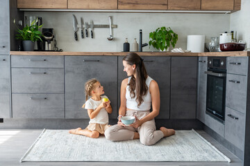 Mother and daughter sitting on kitchen floor, eating or having a breakfast and playing. Family...