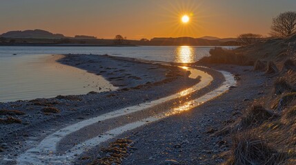 A winding dirt road leads to the water's edge as the sun sets over the horizon, casting a golden glow on the water and the surrounding landscape.