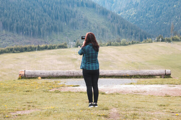 A young girl in a blue green shirt stands in a meadow and takes pictures of mountains covered with pine forest with her mobile phone