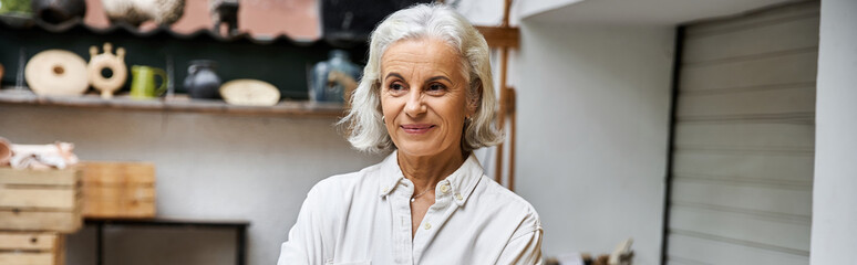A joyful mature woman stands proudly in her art studio surrounded by unique handmade crafts. © Bliss