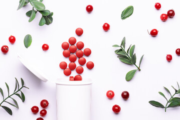 Big red medical tablets in white plastic container on white background with green twigs and raw ripe cranberries close up. Tablets with cranberry extract for UTI treatment. Natural herbal therapy.