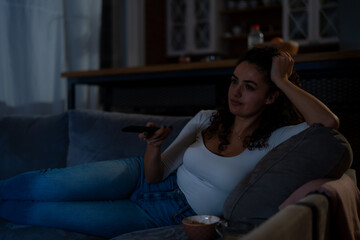 In the evening, a happy curly-haired woman sits comfortably on the couch, enjoying television in a living room lit solely by the TV's glow.