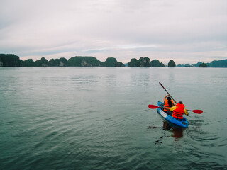 A couple wearing orange lifejackets are kayaking in Halong Bay using paddles going towards the...