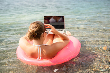 A woman is sitting on a red inflatable raft with a laptop on her lap. She is wearing a white bikini and sunglasses. Concept of relaxation and leisure.