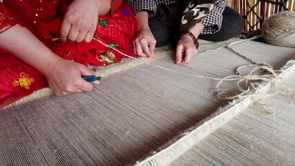 Two women sit on a rug, skillfully weaving a traditional carpet with their hands and small tools