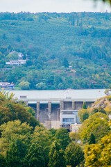 A concrete dam spans a wide reservoir, surrounded by lush greenery. A small village is seen on the opposite shore