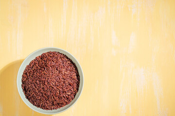 A bowl of red rice is placed on a yellow table.The raw rice is spread out in the bowl, copy-space.