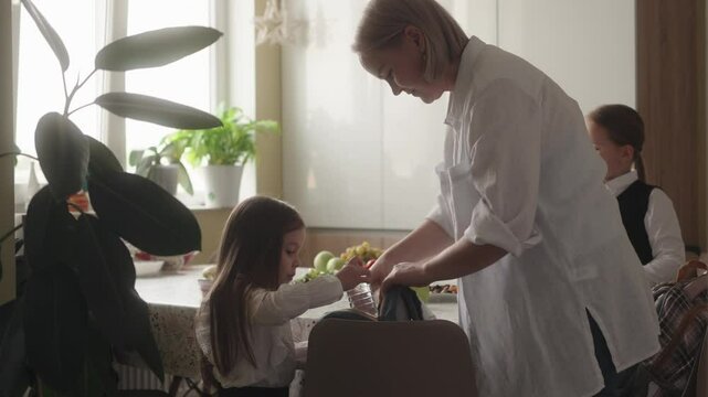 Mother helps her daughters get ready for school by packing snacks and drinks in their lunch bags. The family is preparing for the school day.