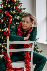 Attractive red-haired, bearded man is sitting on a chair in front of a Christmas tree in a bright living room. Portrait of a serious brutal young guy.New Year and Christmas concept.