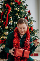 A happy red-bearded young man unpacks Christmas presents at home by the Christmas tree.New Year and Christmas concept.