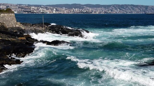 Ocean waves crash against a rocky coast, showcasing the vibrant city of valparaiso in the distance under a sunny sky
