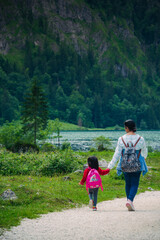 Obraz premium Mother Daughter holding hands and trekking Konigssee Lake in Bavaria Germany with Church by the Lake White Boat Forested Mountain and Beautiful Sunny Trekking in Berchtesgaden German Alps. High