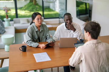 A focused Asian woman and an attentive African man collaborate with a colleague, using a laptop and digital tablet in a bright office environment.