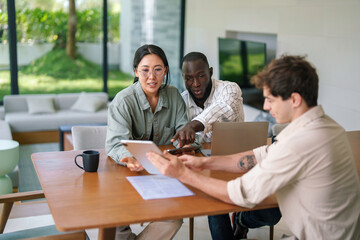 A diverse group of young adult professionals collaborates on a project using tablets and documents in a well-lit modern office.