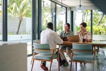 A group of diverse young executives engages in discussion at a round table, using a laptop in a well-lit modern office, conveying a scene of collaborative work and digital interfacing.