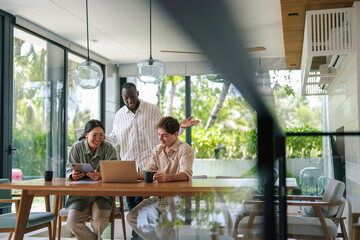 Three young adult business professionals, of varied ethnicities, collaborate using a laptop and digital tablet in a sunlit modern office space.