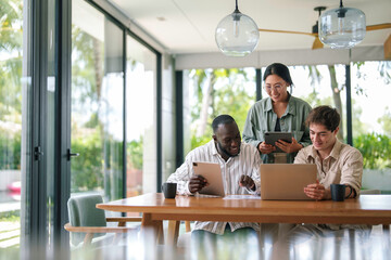 A cheerful group of young diverse business professionals engage in a collaborative project using laptops at a spacious wooden table in a sunlit contemporary office setting.