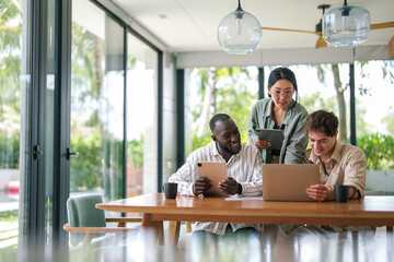A group of cheerful young adults, diverse in ethnicity, engage in a collaborative work session in a bright modern office, using digital tablets and laptops.