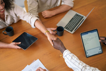 Two young entrepreneurs engage in a friendly handshake over a wooden conference table, surrounded by digital devices and paperwork, signaling the start of a collaborative session in an office space.