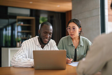 A smiling Black male and Asian female executive work together, analyzing data on a laptop in a well-lit office setting, exemplifying dynamic collaboration.