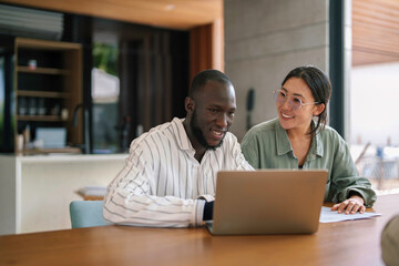 A Black male and Asian female executive work intently at a laptop in a sleek office setting,...